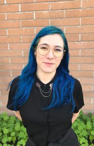 Image of Maeve MacLysaght, fair skinned, blue hair, wearing geometric glasses and a black short-sleeve button-up shirt